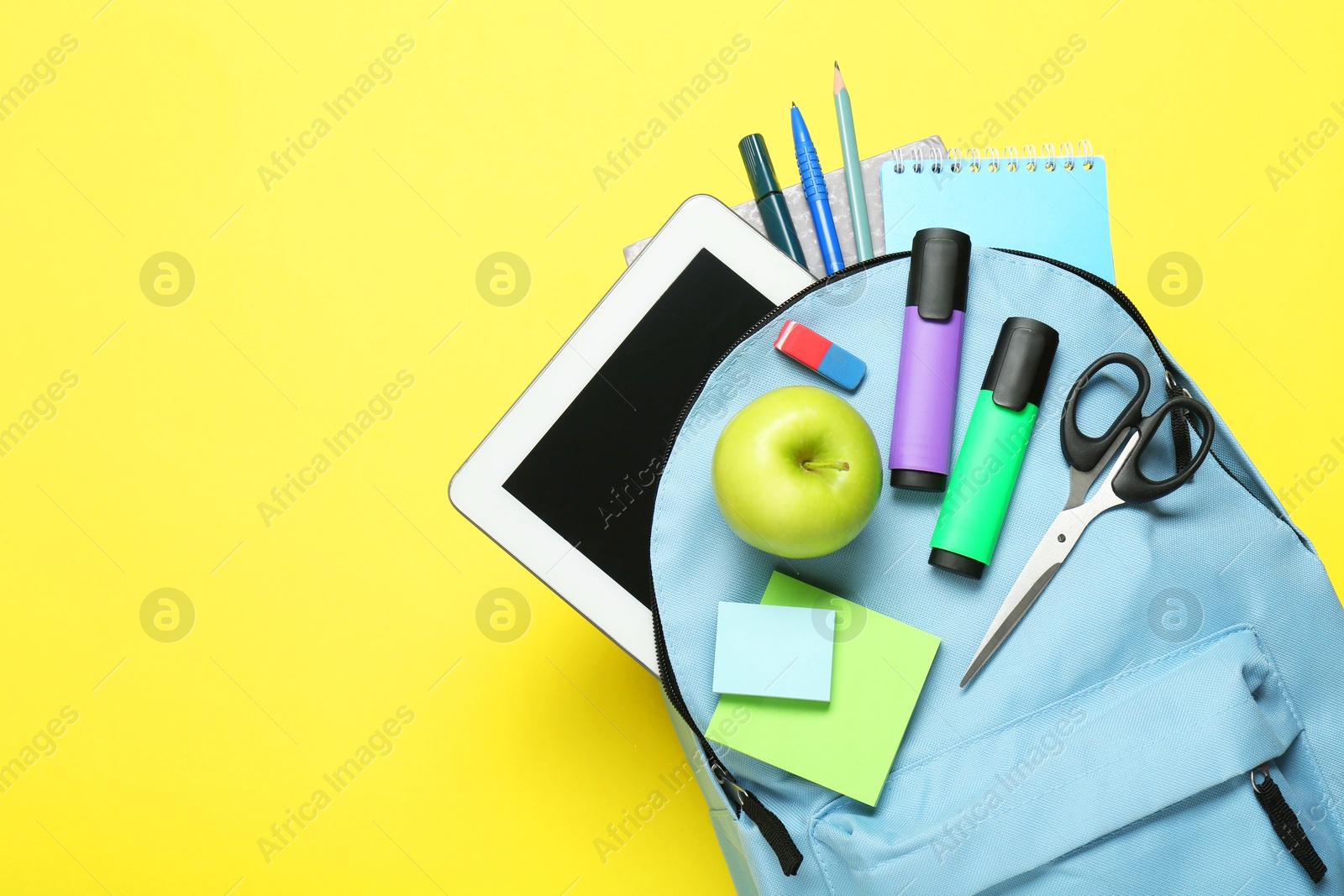 Photo of Light blue school backpack with stationery, apple and tablet on yellow background, top view. Space for text