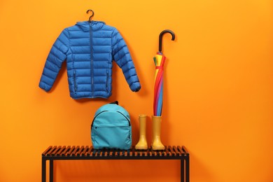 Photo of Colorful umbrella, blue jacket, backpack and rubber boots on wooden bench near orange wall