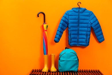 Photo of Colorful umbrella, blue jacket, backpack and rubber boots on wooden bench near orange wall