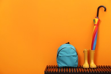 Photo of Colorful umbrella, backpack and rubber boots on wooden bench near orange wall, space for text