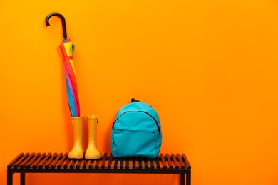 Photo of Colorful umbrella, backpack and rubber boots on wooden bench near orange wall, space for text