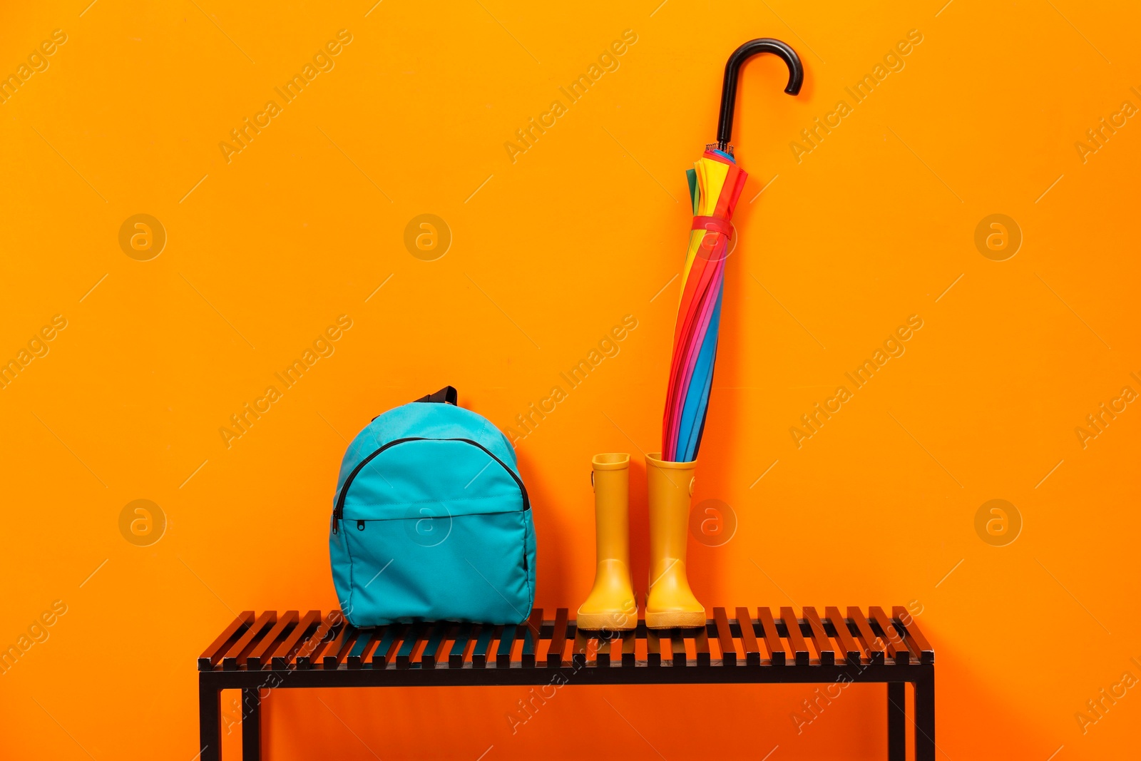 Photo of Colorful umbrella, backpack and rubber boots on wooden bench near orange wall