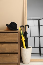 Colorful umbrellas in holder, hat on wooden chest of drawers and mirror indoors