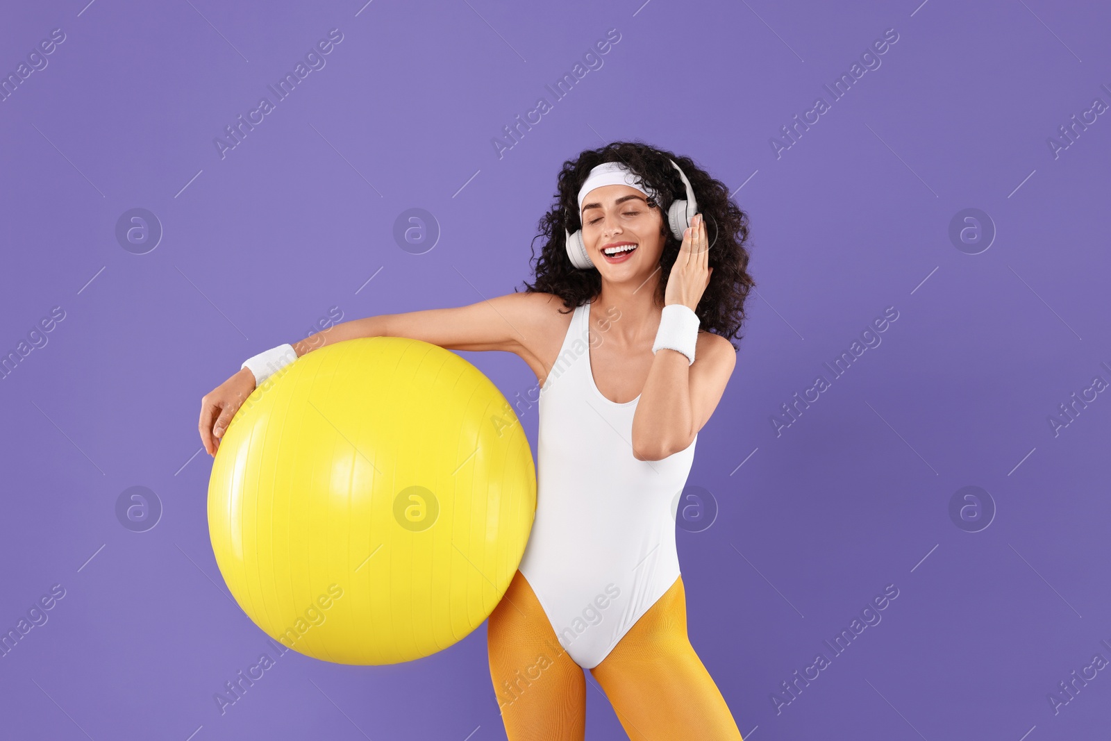 Photo of Aerobics. Happy woman in headphones with fitness ball on purple background