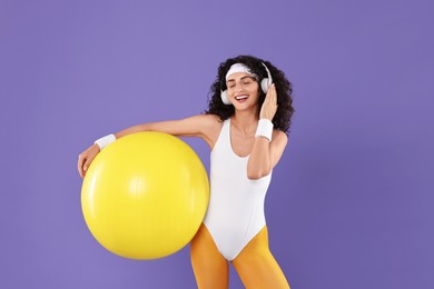 Photo of Aerobics. Happy woman in headphones with fitness ball on purple background