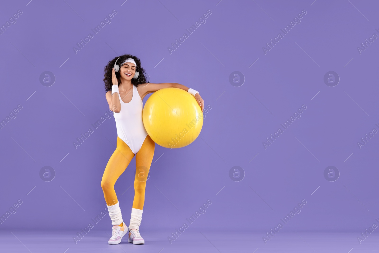 Photo of Aerobics. Happy woman in headphones with fitness ball on purple background, space for text