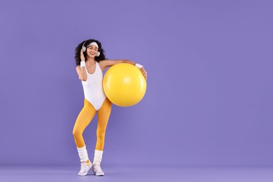 Aerobics. Happy woman in headphones with fitness ball on purple background, space for text