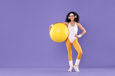 Photo of Aerobics. Happy woman in headphones with fitness ball on purple background, space for text