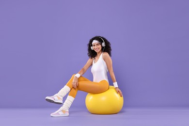 Aerobics. Happy woman in headphones with fitness ball on purple background