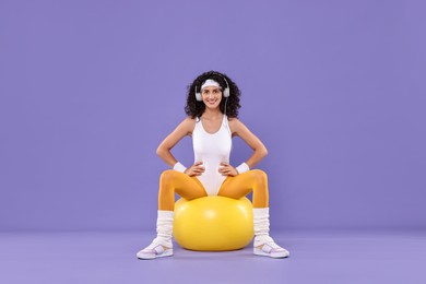 Photo of Woman in headphones doing aerobic exercise with dumbbells and fitness ball on purple background