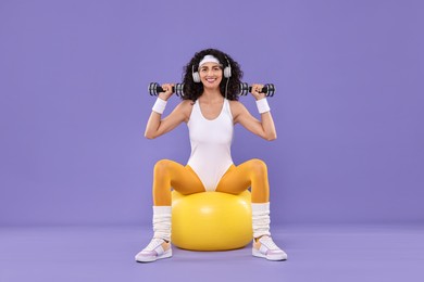 Photo of Woman in headphones doing aerobic exercise with dumbbells and fitness ball on purple background