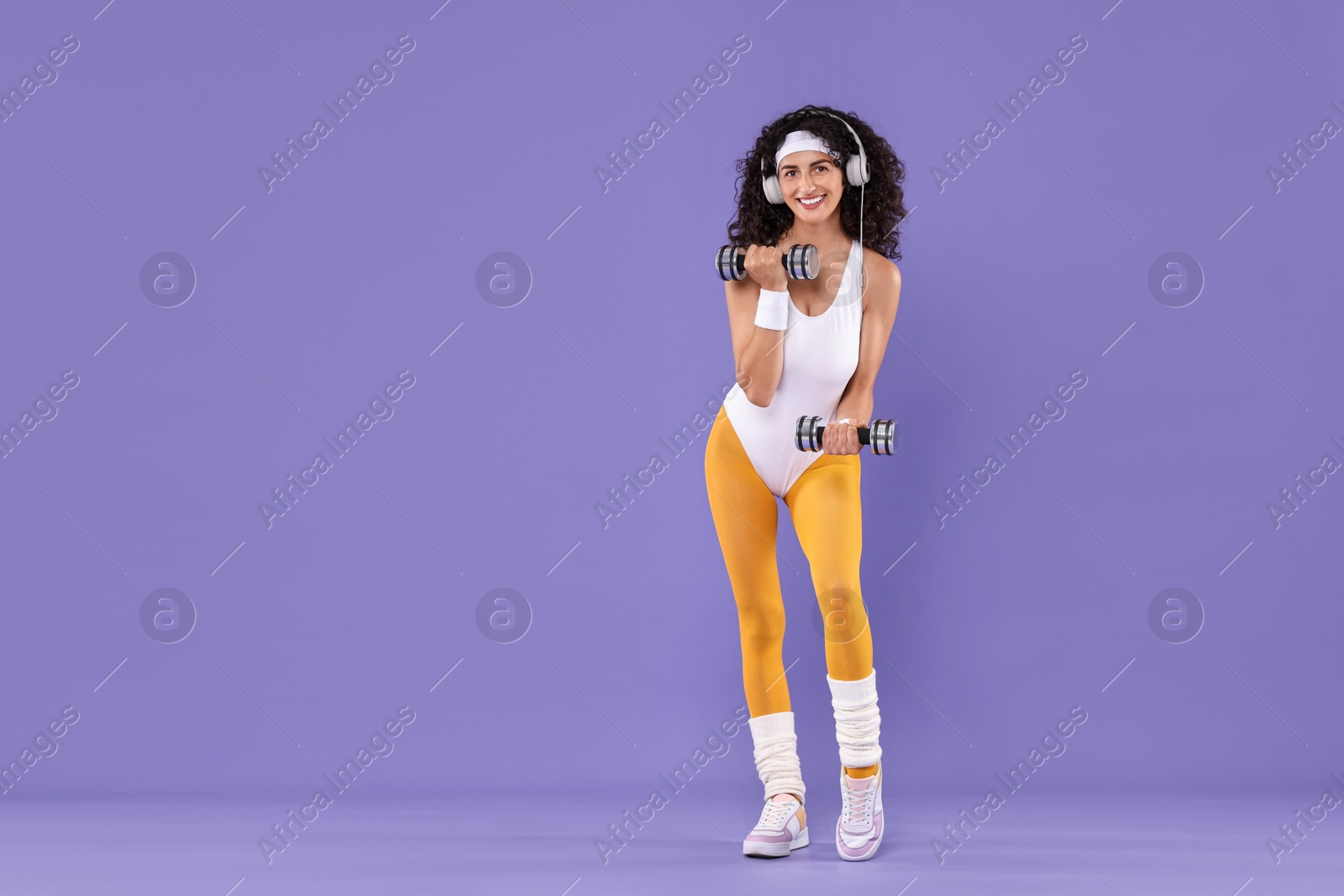 Photo of Woman in headphones doing aerobic exercise with dumbbells on purple background. Space for text