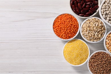 Photo of Different types of cereals and legumes in bowls on white wooden table, flat lay. Space for text