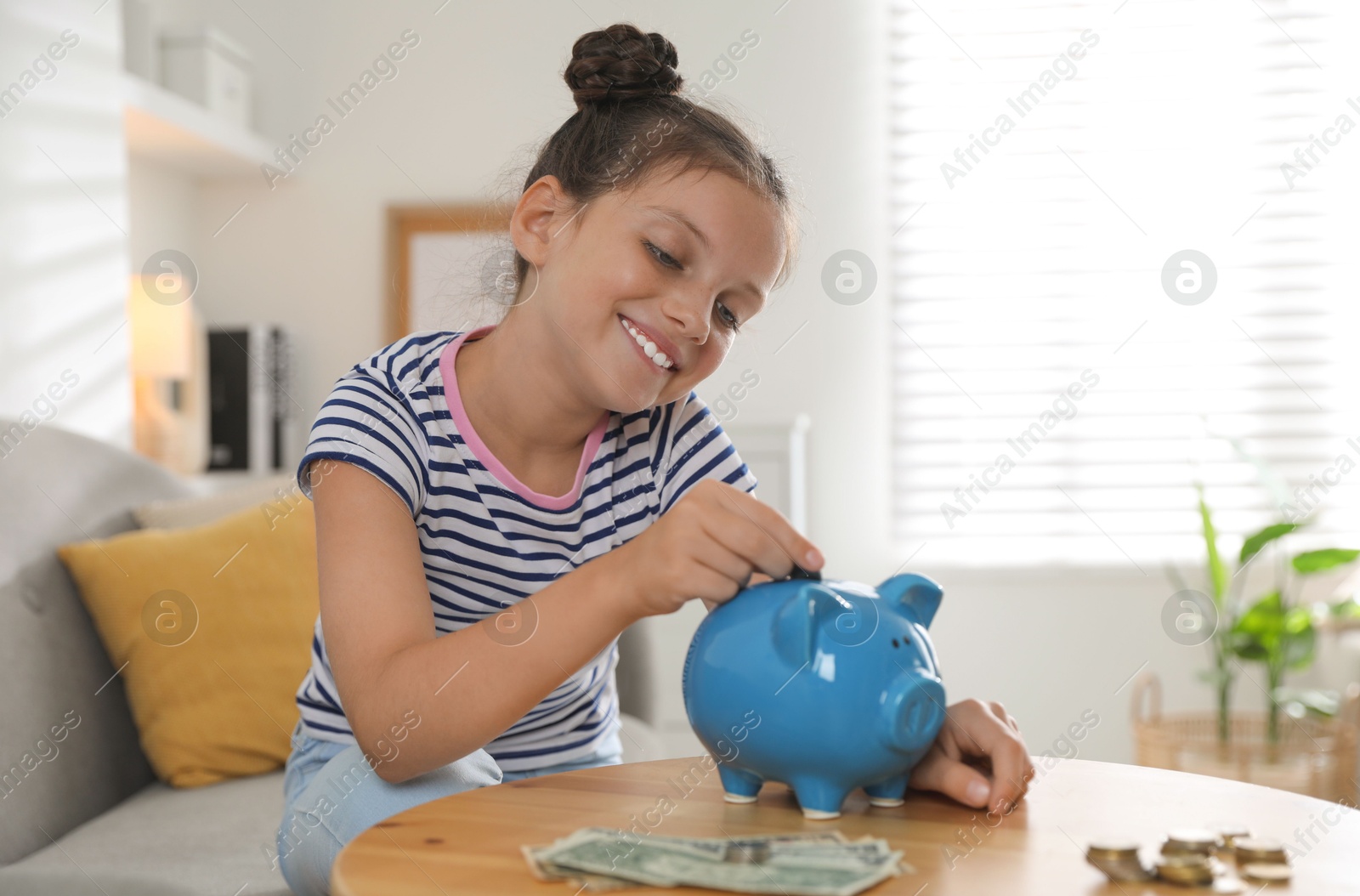 Photo of Pocket money. Cute girl putting coin into piggy bank at wooden table indoors