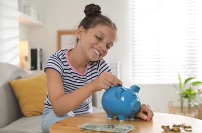 Photo of Pocket money. Cute girl putting coin into piggy bank at wooden table indoors