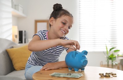 Photo of Pocket money. Cute girl putting coin into piggy bank at wooden table indoors