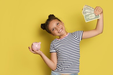 Photo of Pocket money. Cute girl with piggy bank and dollar banknotes on yellow background