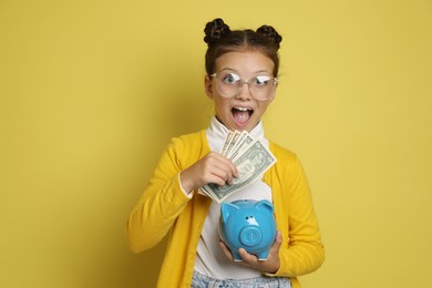 Photo of Pocket money. Cute girl with piggy bank and dollar banknotes on yellow background