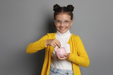 Photo of Pocket money. Cute girl putting coins into piggy bank on grey background