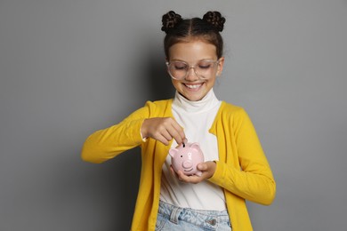 Pocket money. Cute girl putting coins into piggy bank on grey background