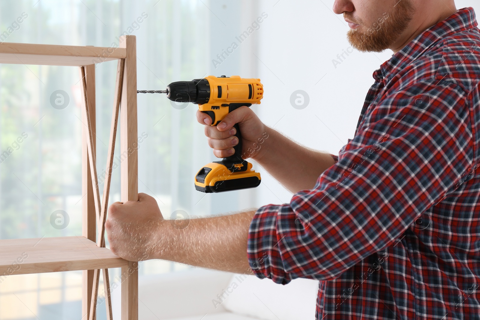 Photo of Man with electric screwdriver assembling wooden shelving unit in room