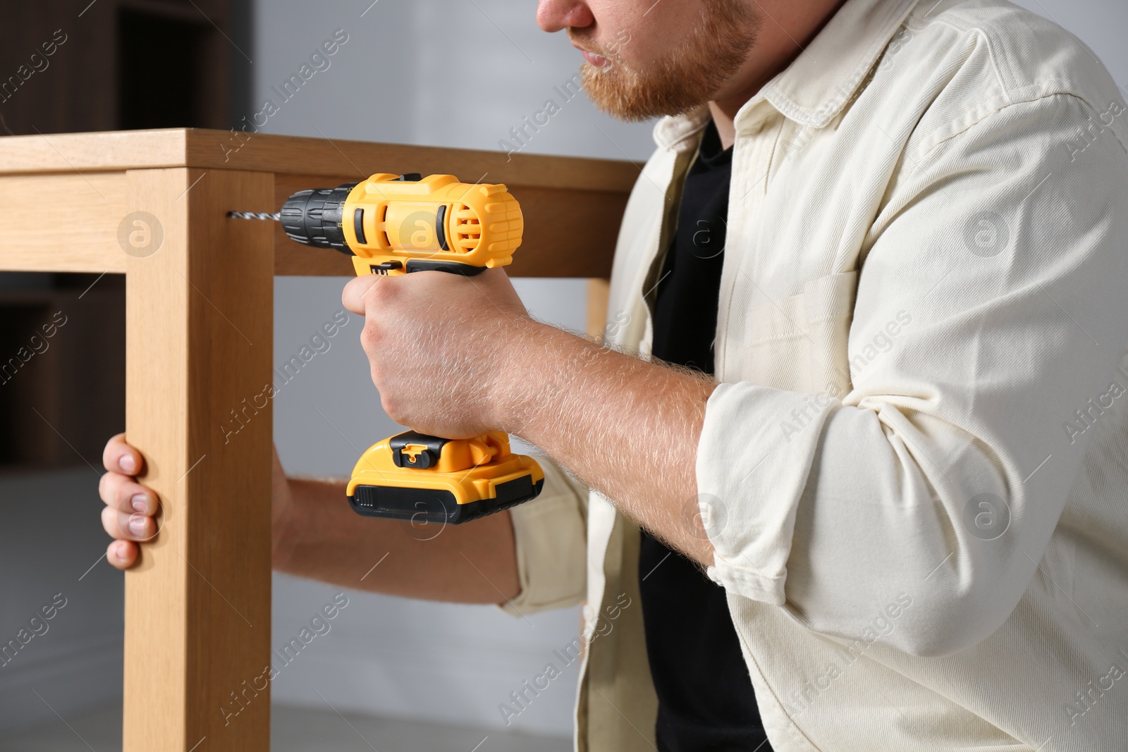 Photo of Man with electric screwdriver assembling table indoors, closeup