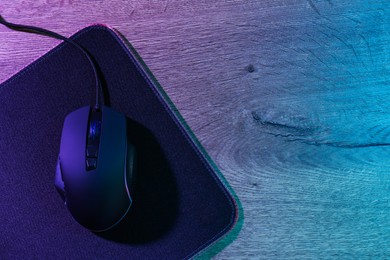 Photo of Black computer mouse and mousepad on wooden table in neon lights, top view. Space for text