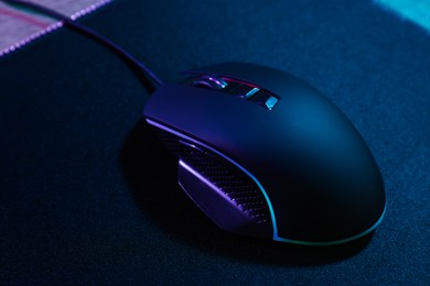 Photo of Black computer mouse and mousepad on wooden table in neon lights, closeup