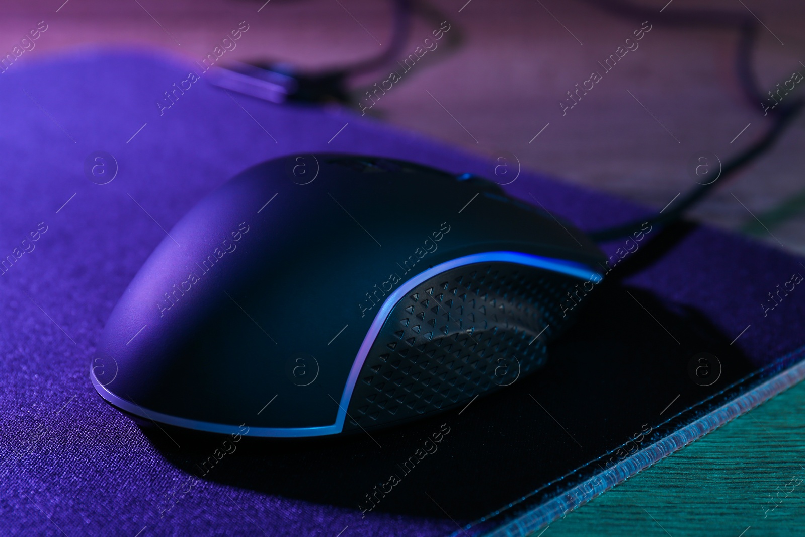 Photo of Black computer mouse and mousepad on wooden table in neon lights, closeup