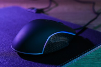 Photo of Black computer mouse and mousepad on wooden table in neon lights, closeup