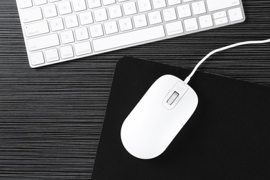 Photo of White computer mouse with mousepad and keyboard on black wooden table, flat lay