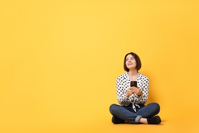 Photo of Smiling woman with smartphone sitting on yellow background. Space for text