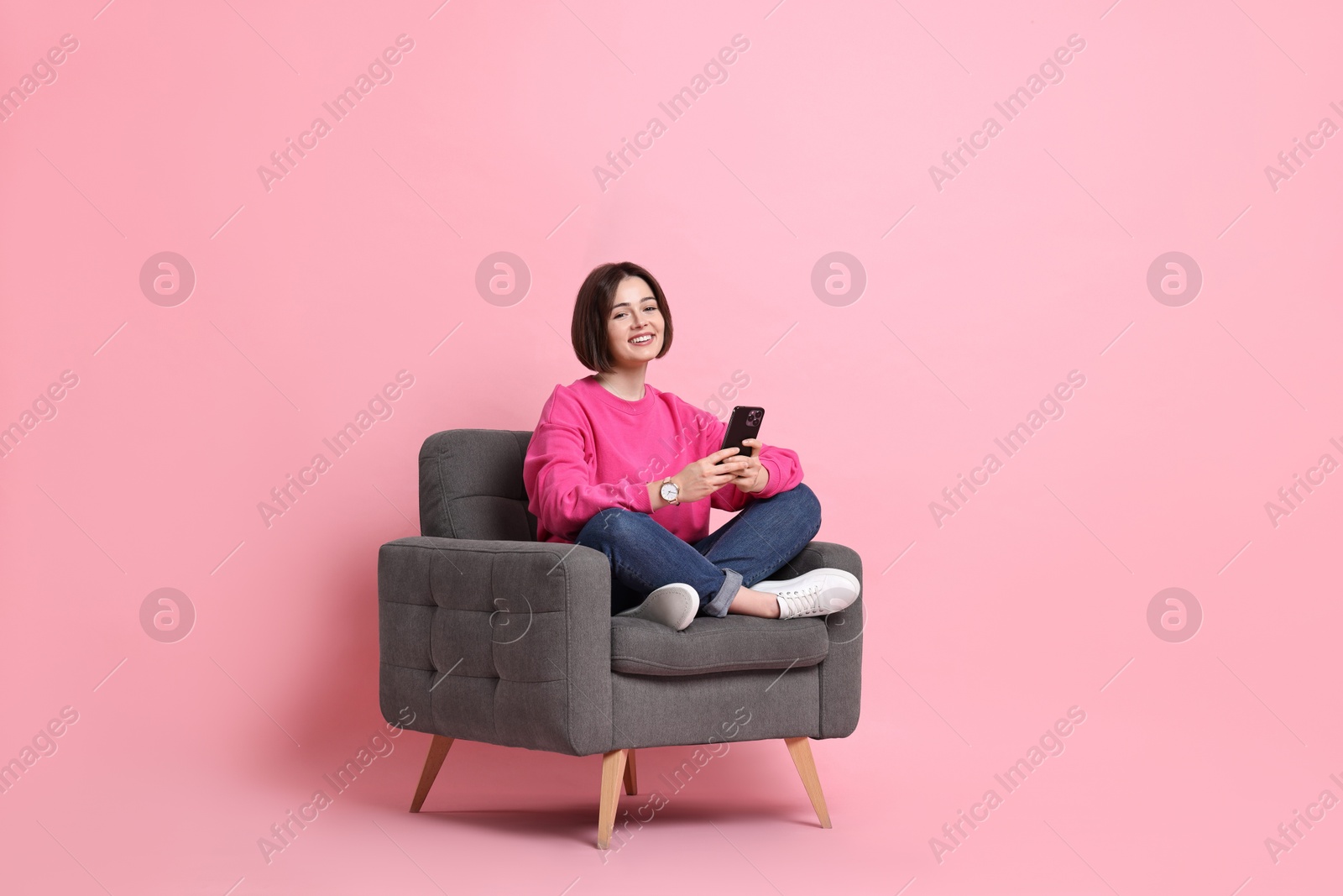 Photo of Smiling woman with smartphone sitting on armchair against pink background