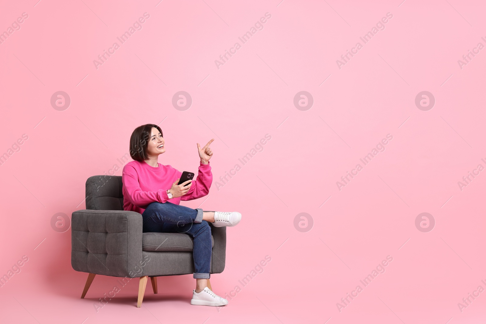 Photo of Smiling woman with smartphone pointing at something against pink background. Space for text