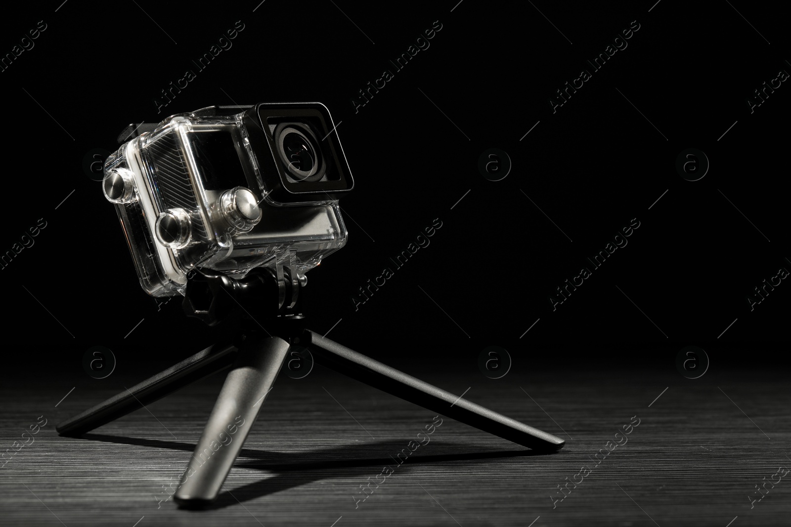 Photo of Modern action camera and tripod on wooden surface against black background. Space for text