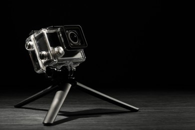 Modern action camera and tripod on wooden surface against black background. Space for text