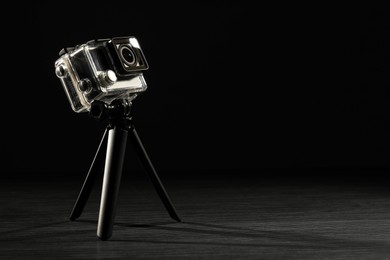 Modern action camera and tripod on wooden surface against black background. Space for text