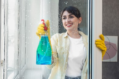 Beautiful young woman with spray bottle of detergent cleaning window indoors