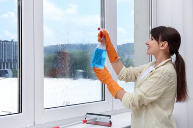 Beautiful young woman with spray bottle of detergent cleaning window indoors