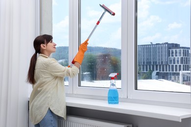 Beautiful young woman with squeegee tool cleaning window indoors