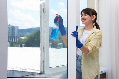 Beautiful young woman with spray bottle of detergent cleaning window indoors