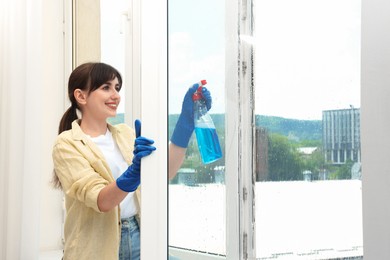 Beautiful young woman with spray bottle of detergent cleaning window indoors