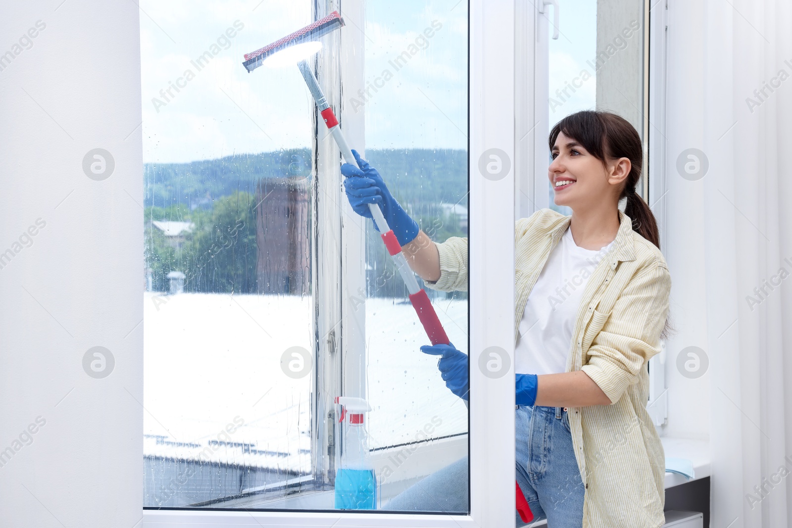 Photo of Beautiful young woman with squeegee tool cleaning window indoors