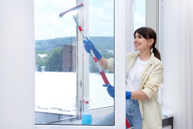 Photo of Beautiful young woman with squeegee tool cleaning window indoors