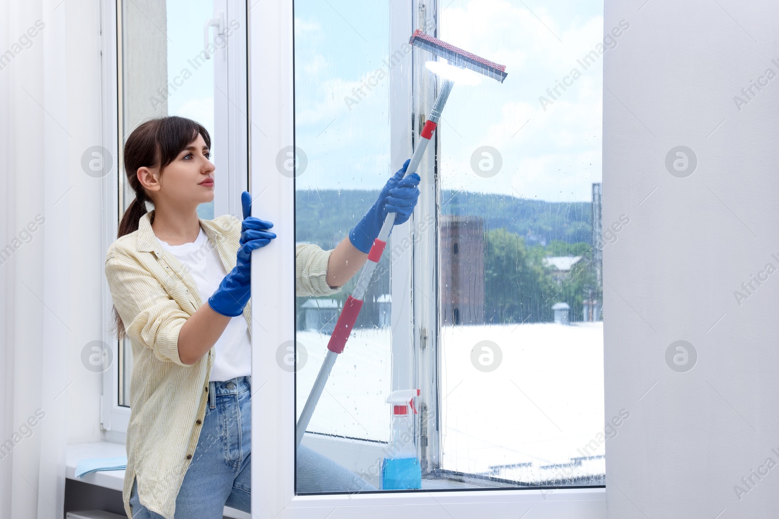 Photo of Beautiful young woman with squeegee tool cleaning window indoors