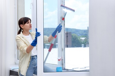 Photo of Beautiful young woman with squeegee tool cleaning window indoors