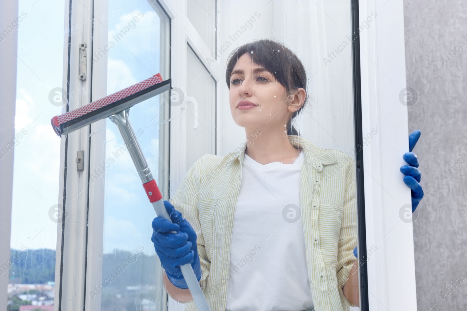 Photo of Beautiful young woman with squeegee tool cleaning window indoors