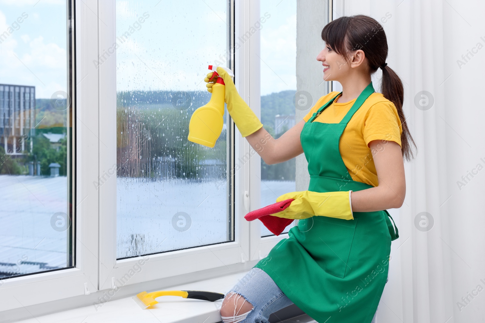 Photo of Housewife with spray bottle of detergent and napkin cleaning window indoors