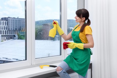 Photo of Housewife with spray bottle of detergent and napkin cleaning window indoors