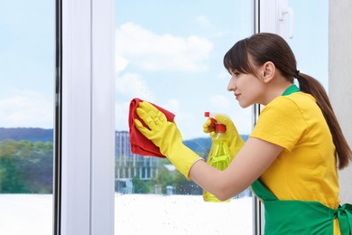 Housewife with spray bottle of detergent and napkin cleaning window indoors